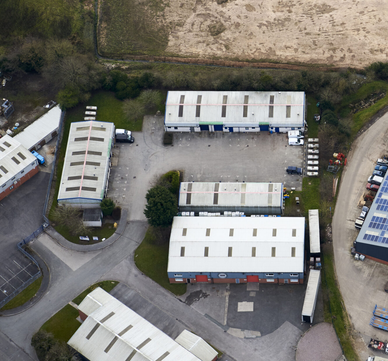 Aerial view of an industrial park with several rectangular warehouse buildings, parking areas, and a few vehicles on paved roads surrounded by grassy land.