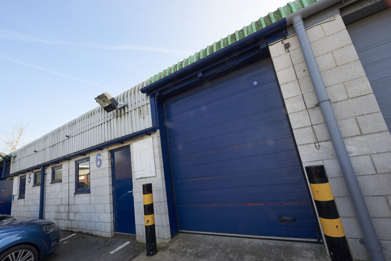 A blue industrial garage door with the number 6 on a concrete building, security camera above, and yellow-black striped bollards in front.