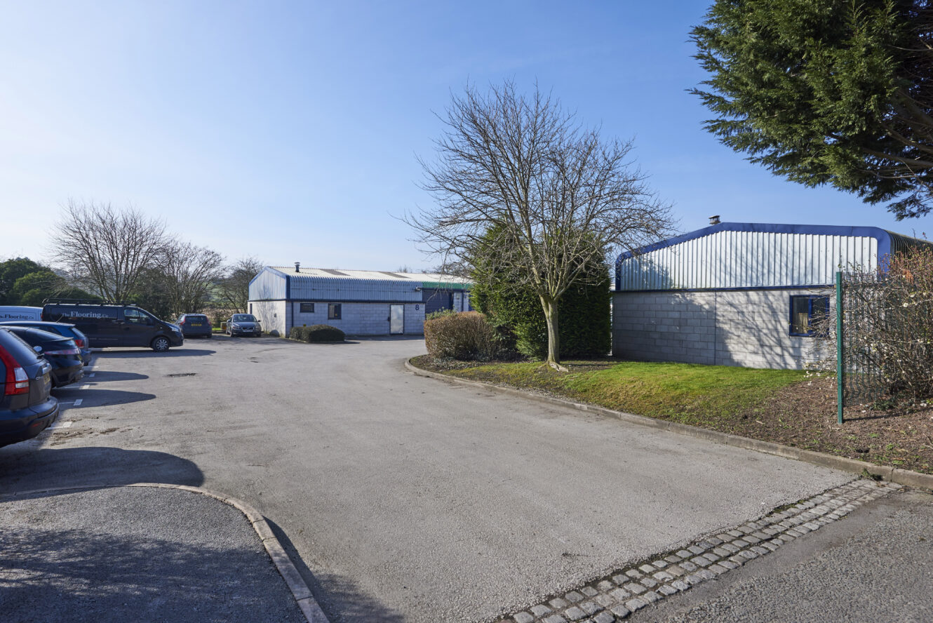 A paved parking lot with several cars and vans, bordered by industrial buildings, trees, and shrubs on a clear, sunny day.