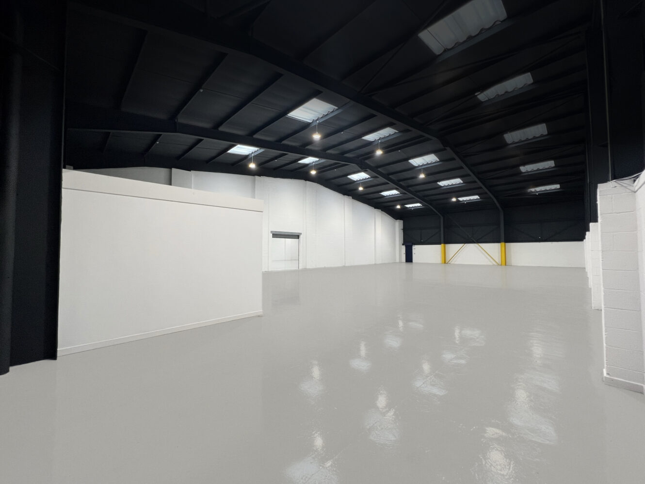 Spacious, empty industrial warehouse with a polished gray floor, high black ceiling, and white walls, illuminated by overhead lights.