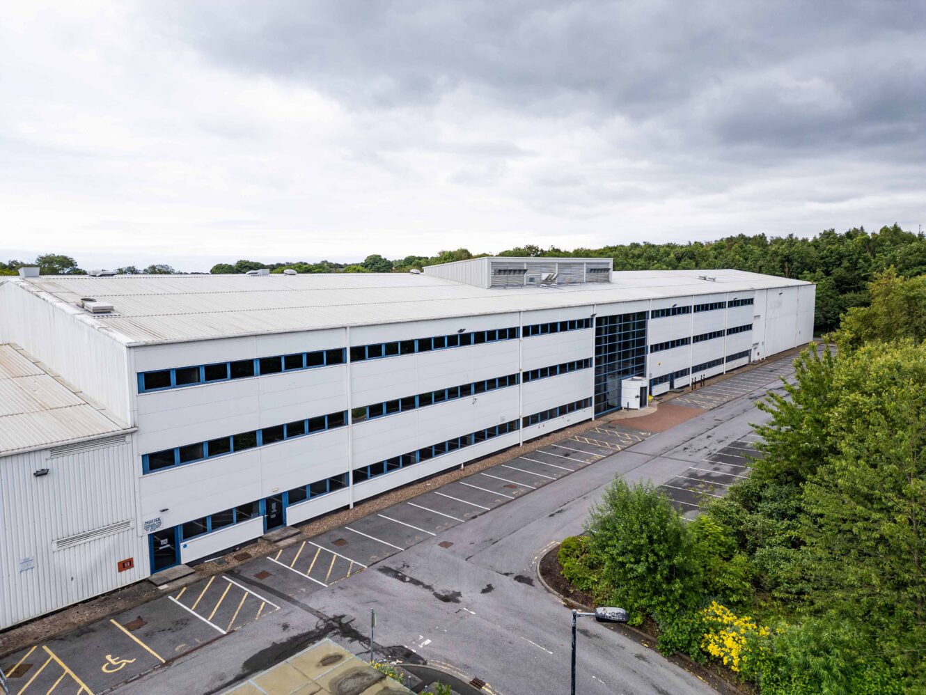 Large, white, two-story industrial building with blue-trimmed windows, surrounded by trees and an empty parking lot under a cloudy sky.