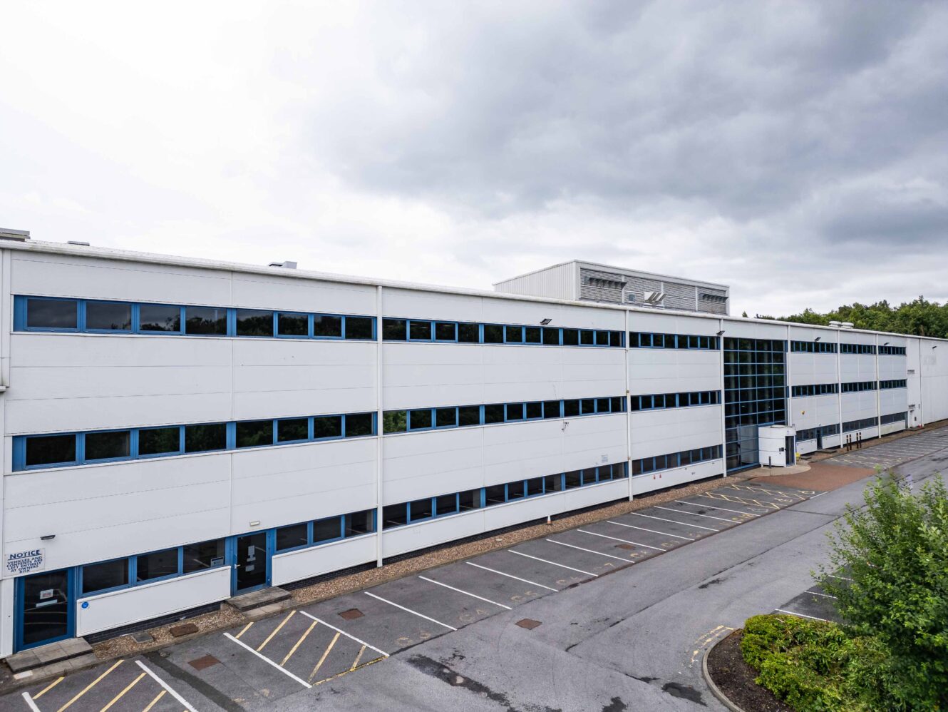 A large, modern, white office or industrial building with blue windows, an empty parking lot, and cloudy sky overhead.