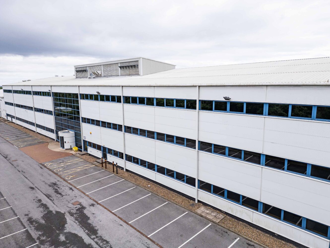A large, modern, white industrial or office building with blue window frames and an empty adjacent parking lot under a cloudy sky.