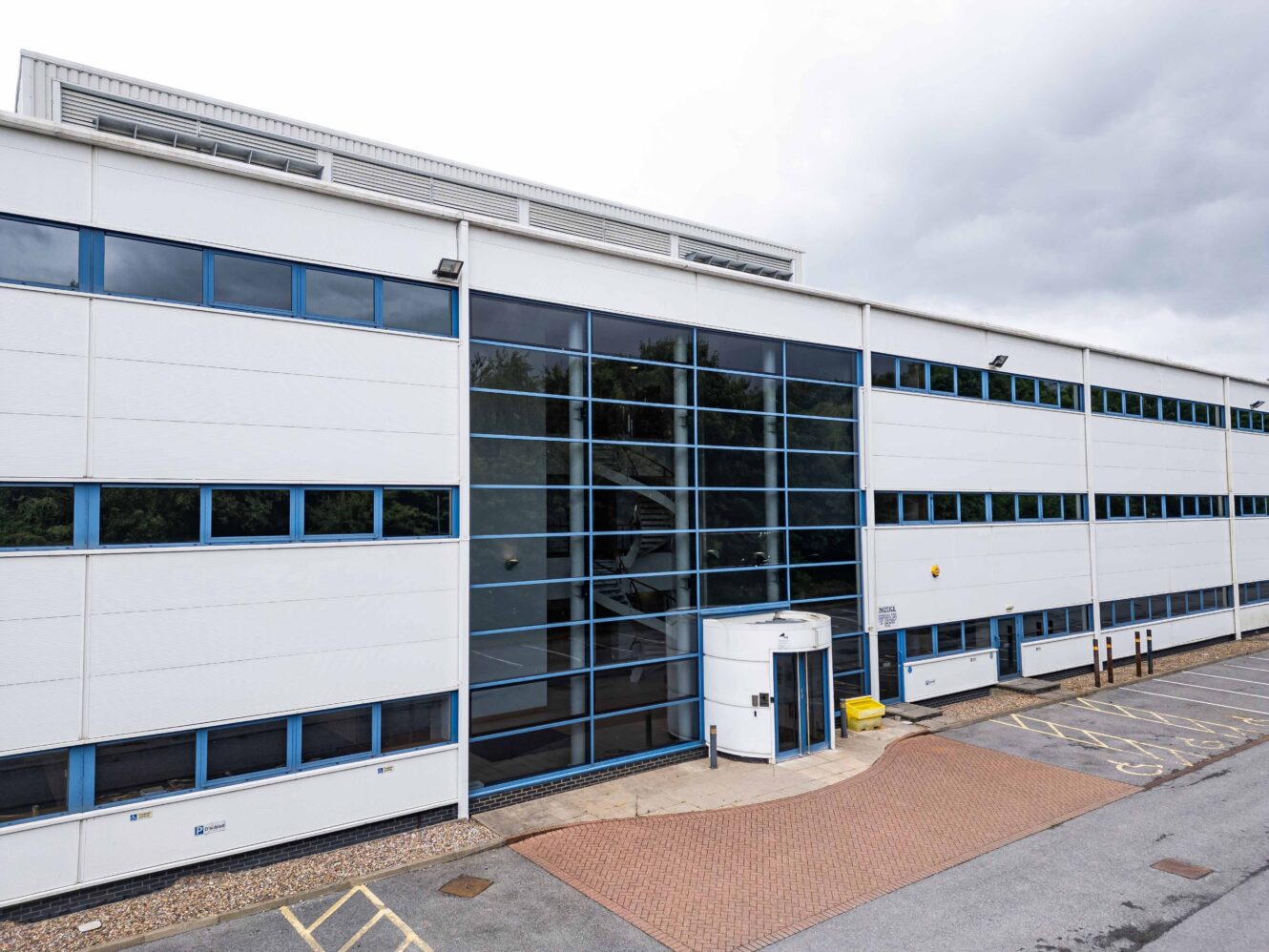 Modern white commercial or office building with large glass windows, accessible entrance, and adjacent disabled parking spaces on a cloudy day.
