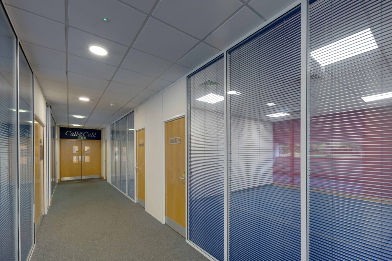 A hallway with carpeted floor, glass office partitions with blinds, wooden doors, and fluorescent ceiling lights, leading to a door labeled Café.