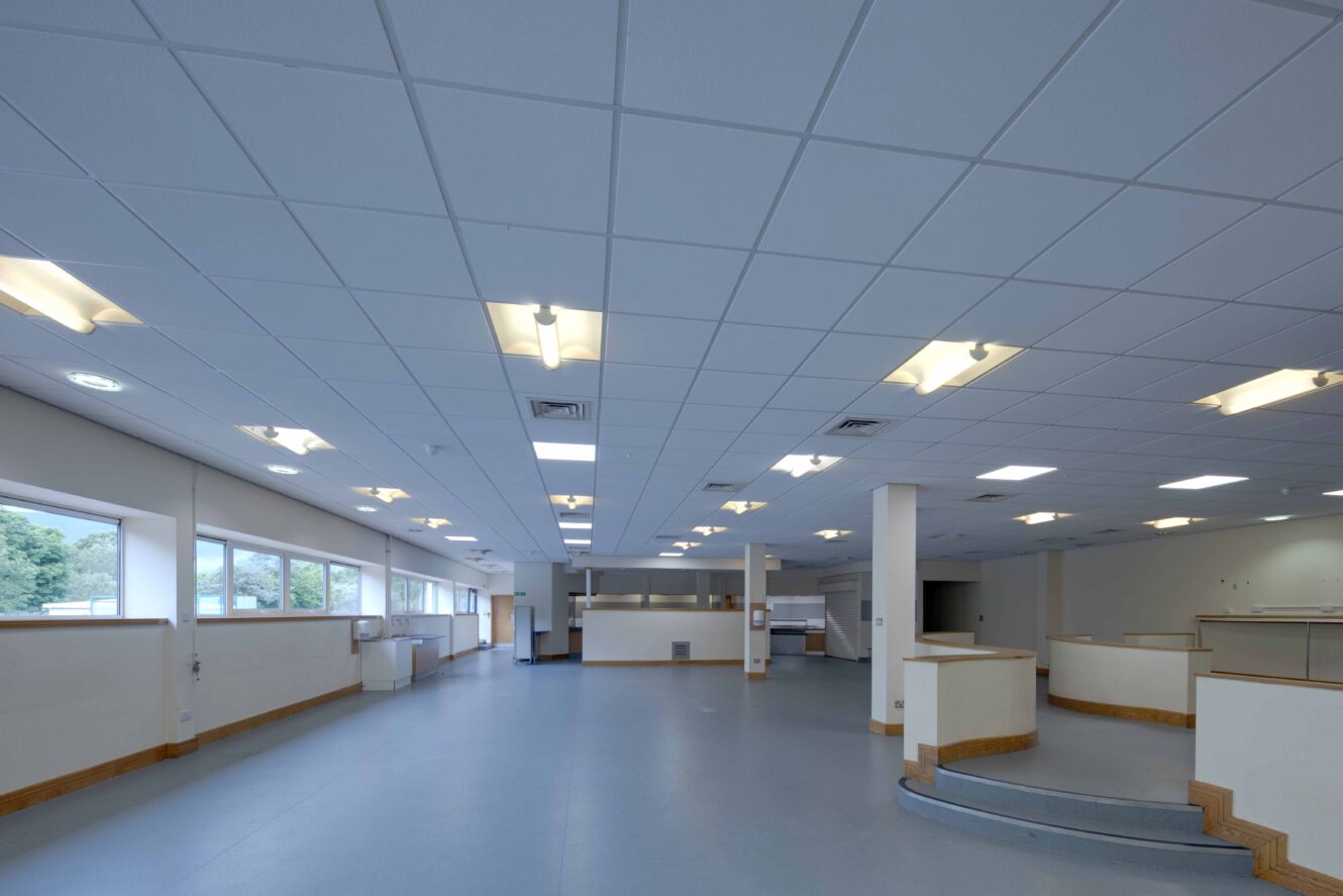 Spacious, empty room with blue flooring, white walls, ceiling lights, large windows on one side, and partitioned workspaces.