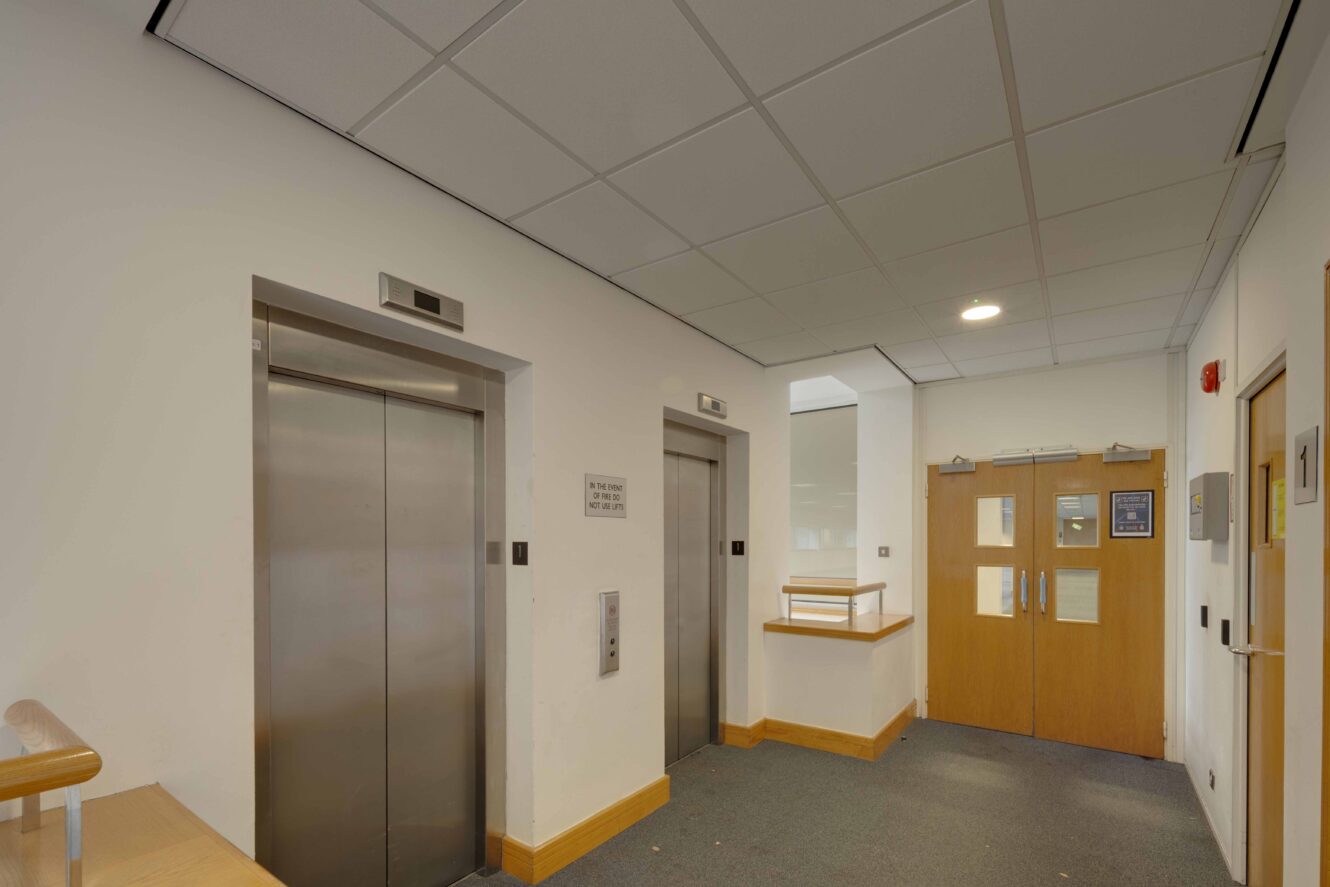 A hallway with two closed elevators on the left and double wooden doors with glass panels on the right. The floor is carpeted and the ceiling has recessed lighting.