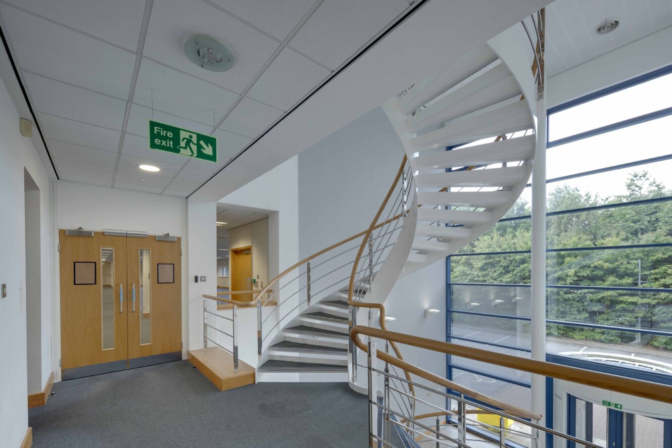 Modern interior stairwell with white steps, wooden handrails, large windows, double doors, and a green fire exit sign on the ceiling.