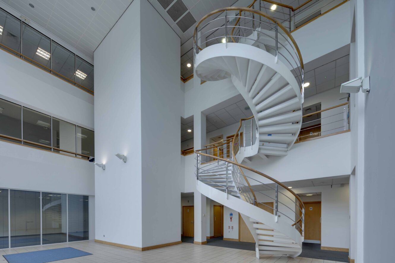 Modern building interior with a white spiral staircase, tiled floor, glass walls, and multiple stories visible.
