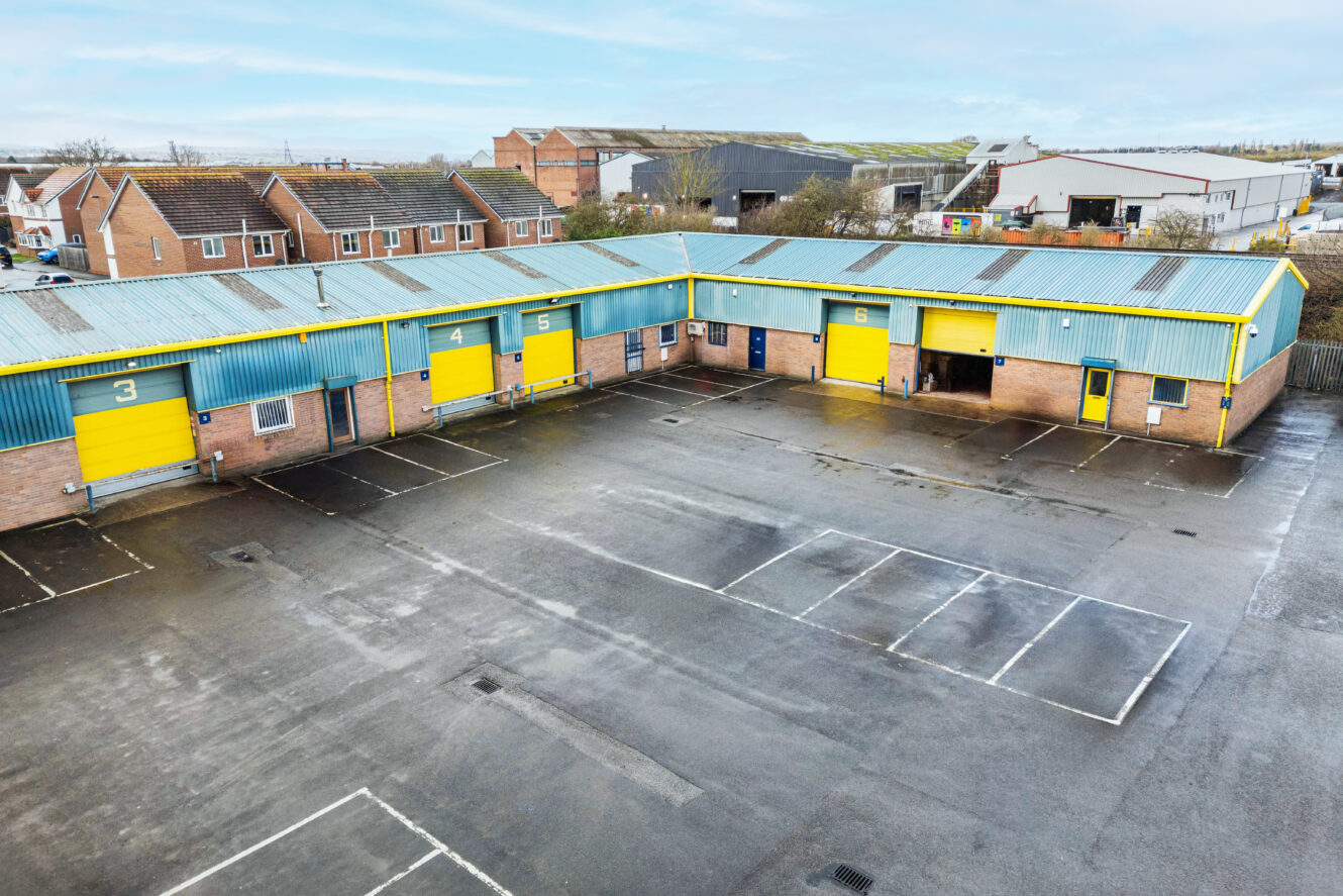 Industrial units with yellow and blue doors, numbered 3, 4, 5, and 6, in a row next to an empty parking lot and nearby residential houses.