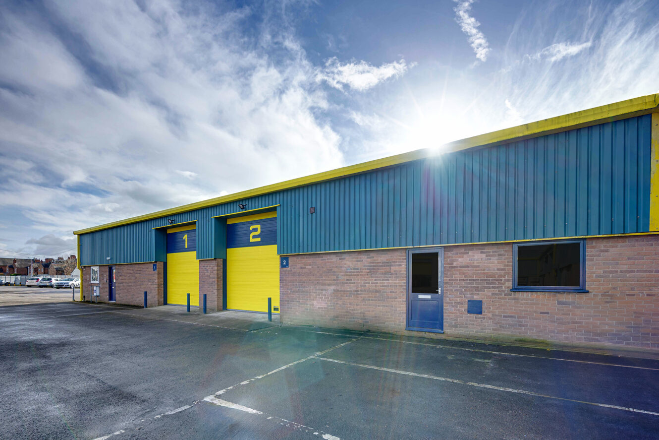 A row of industrial warehouse units with blue and yellow doors numbered 1 and 2, under a partly cloudy sky with sunlight in the background.