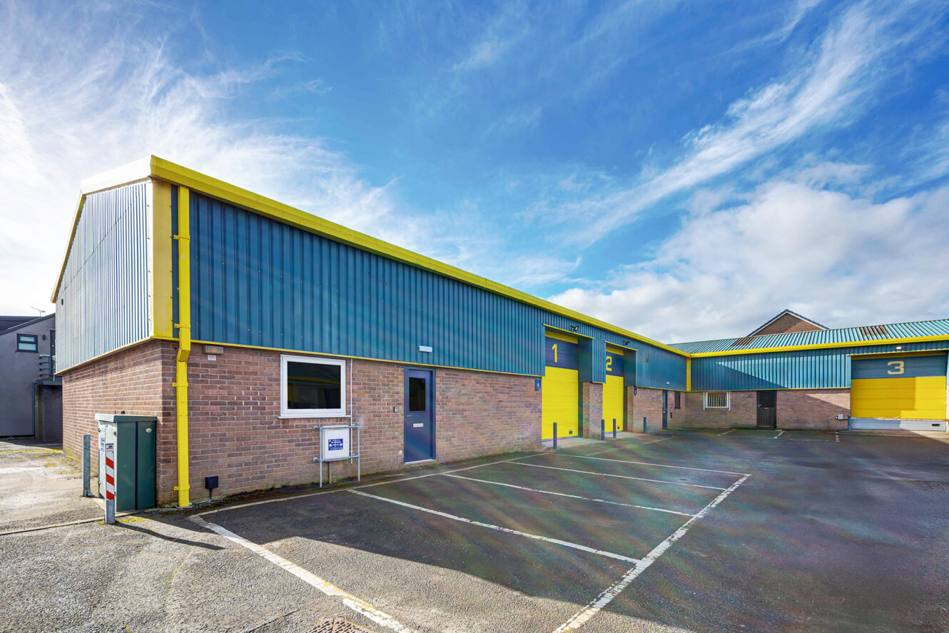 A row of industrial warehouse units with blue and yellow exteriors, numbered doors, and an empty parking lot under a partly cloudy sky.