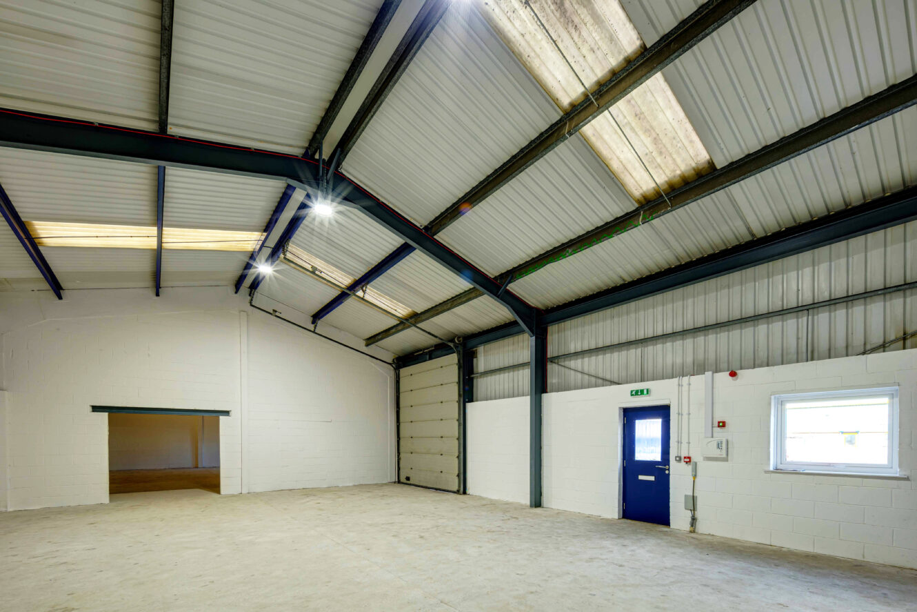 Empty industrial warehouse with a high metal ceiling, concrete floor, white walls, blue door, window, and overhead lights.