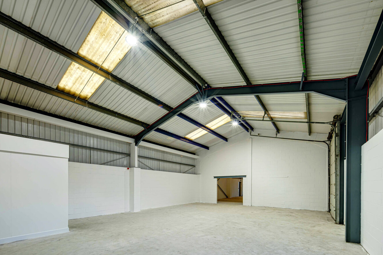 Spacious empty warehouse interior with high ceilings, metal beams, white brick walls, and large entryway under bright overhead lighting.