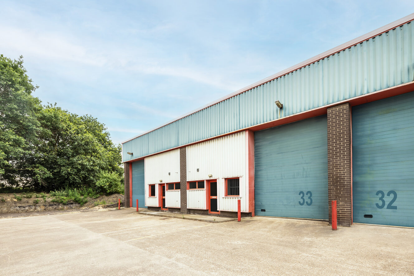 Exterior of an industrial warehouse with numbered blue roller doors 32 and 33, small entry doors, and a paved lot, next to trees under a partly cloudy sky.