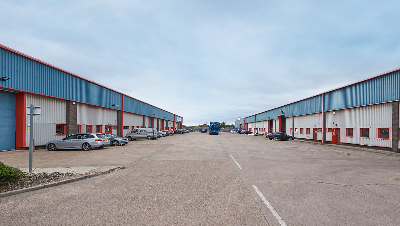 A wide concrete lot between two large industrial warehouse buildings with parked cars along both sides and a cloudy sky overhead.