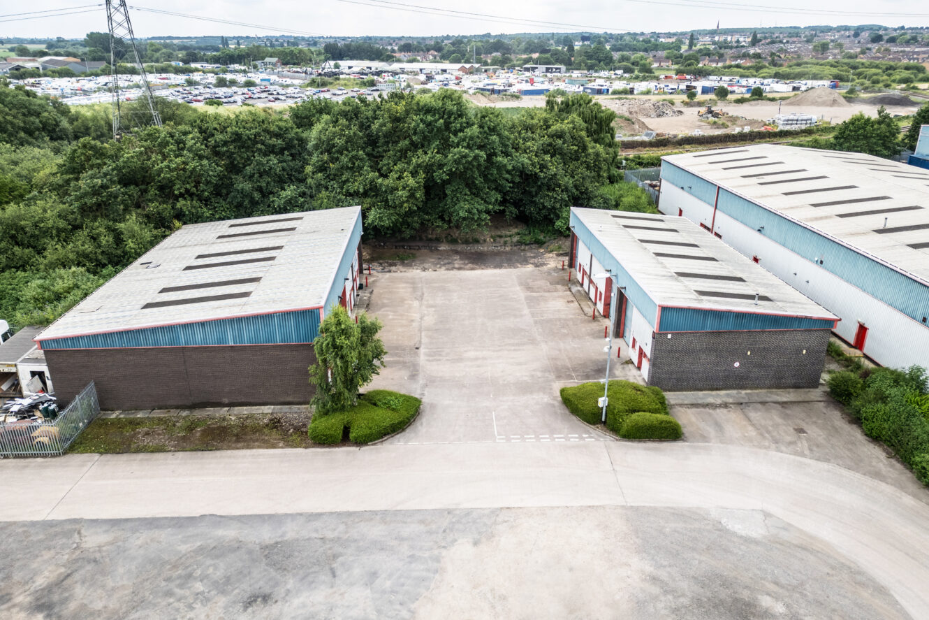 Aerial view of two industrial warehouse buildings separated by a concrete yard, surrounded by trees and greenery, with a parking lot and landscape in the background.