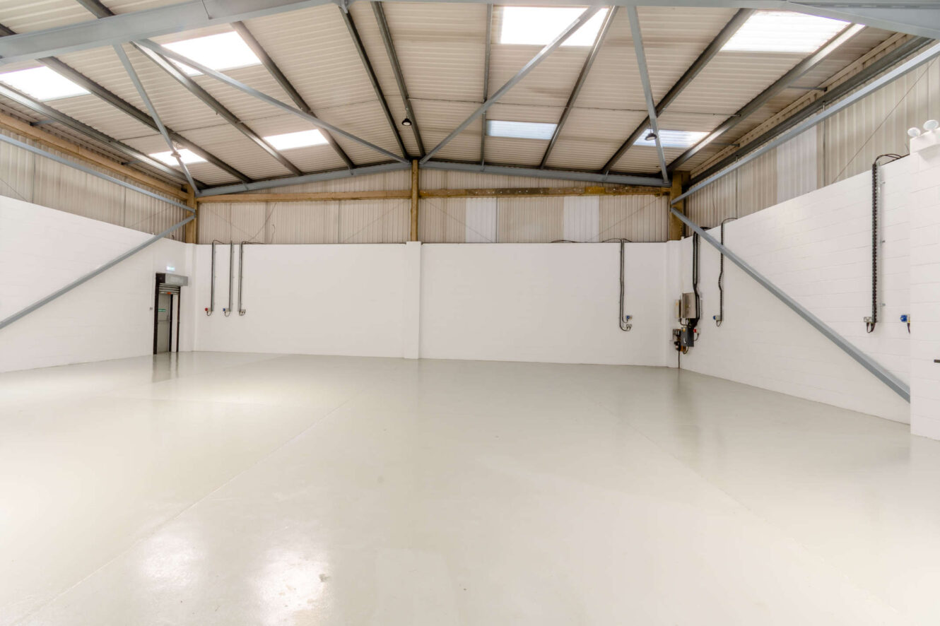 Large, empty industrial warehouse with white walls, high ceiling, exposed beams, and several utility conduits along the walls.