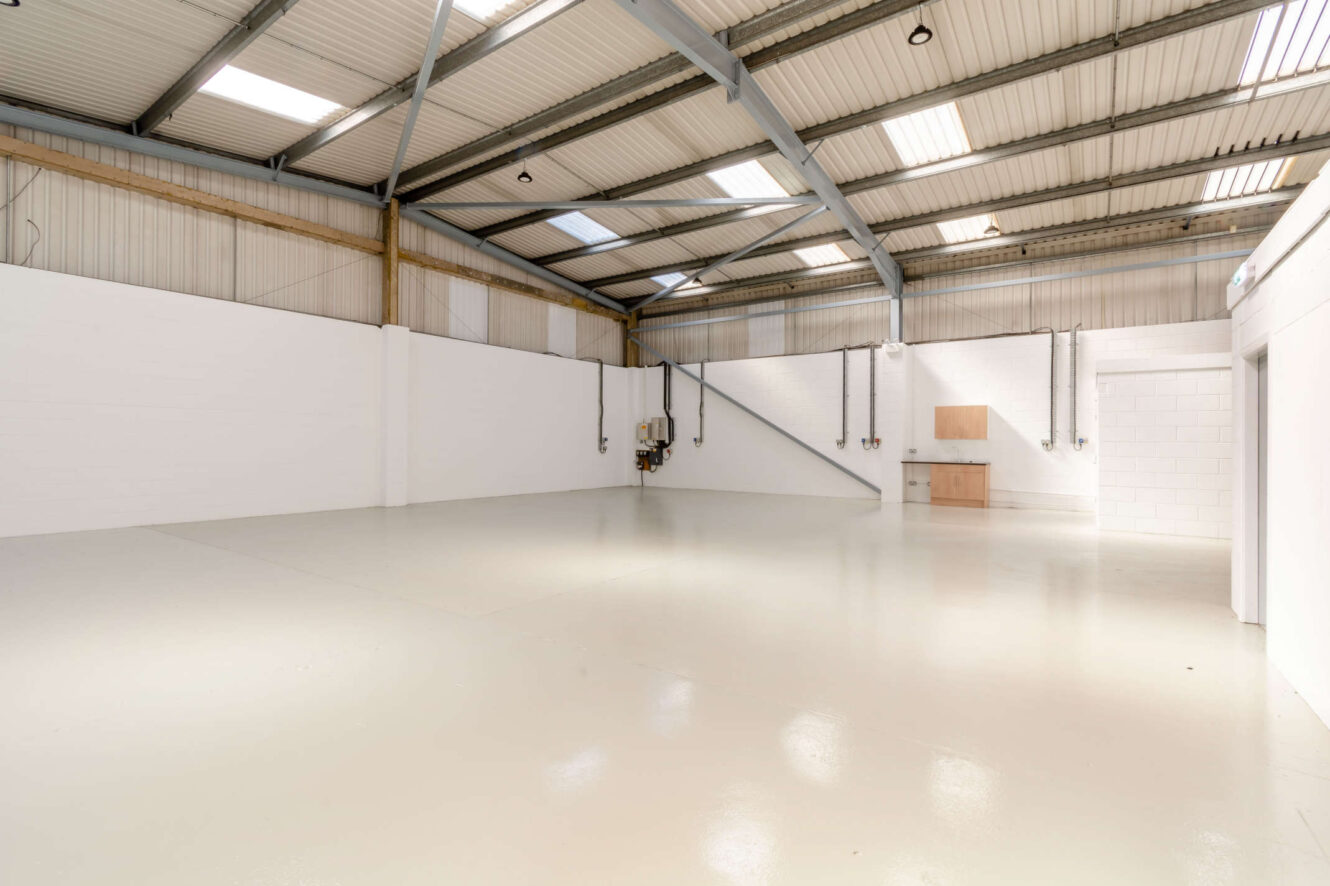 Empty industrial warehouse space with white walls, high metal ceiling, exposed beams, fluorescent lighting, and a small utility area with a sink and cabinets in the corner.