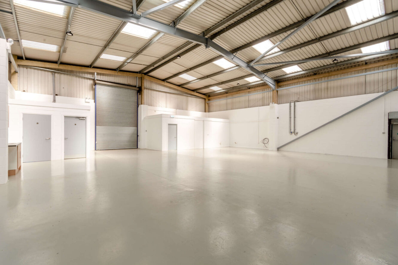 Spacious empty warehouse with high ceilings, white walls, concrete floor, large roller door, and overhead lighting.