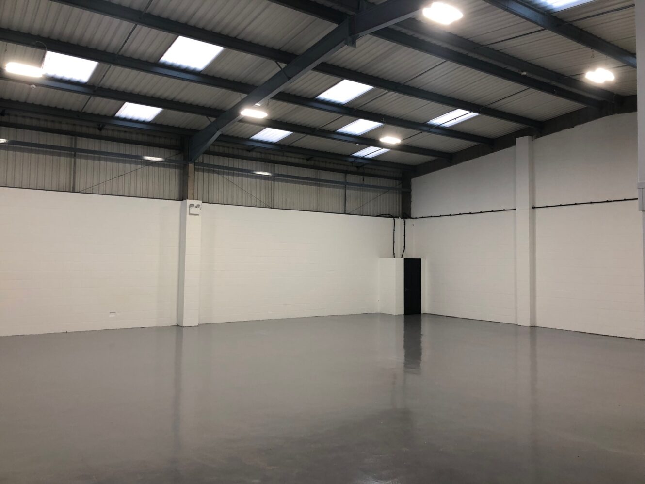 Empty industrial warehouse with gray polished floor, white painted brick walls, high ceiling, and large overhead lights. A black door is visible in the corner.