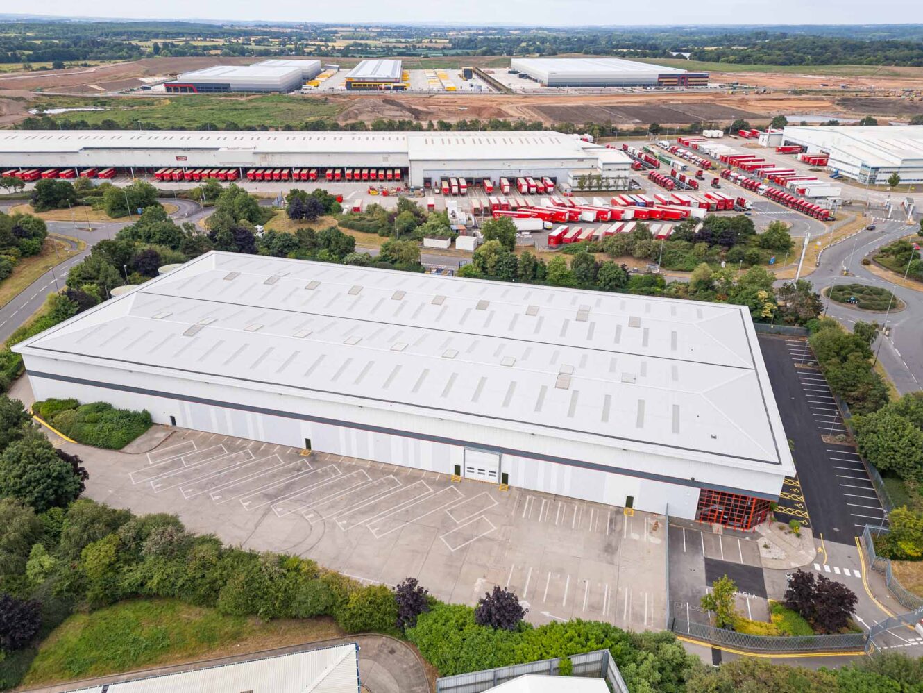 A large industrial warehouse with an empty parking lot, surrounded by trees and roads, with other warehouses and red vehicles in the background.