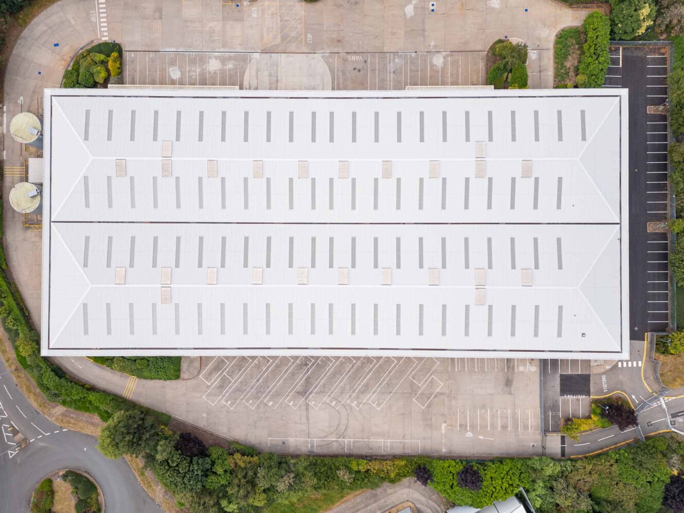 Aerial view of a large rectangular warehouse with a white roof, surrounded by parking lots, greenery, and access roads.