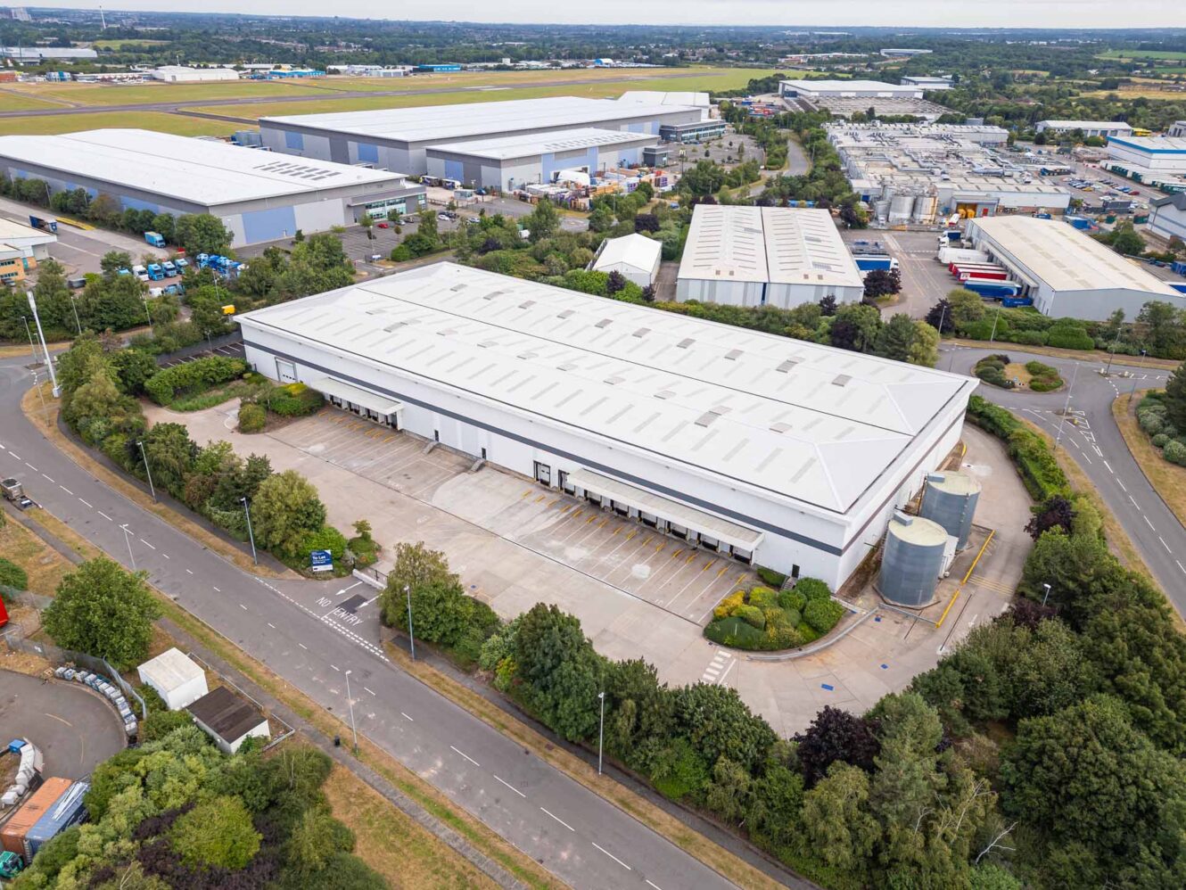 Aerial view of a large white warehouse with loading bays, surrounded by roads, greenery, and other industrial buildings.