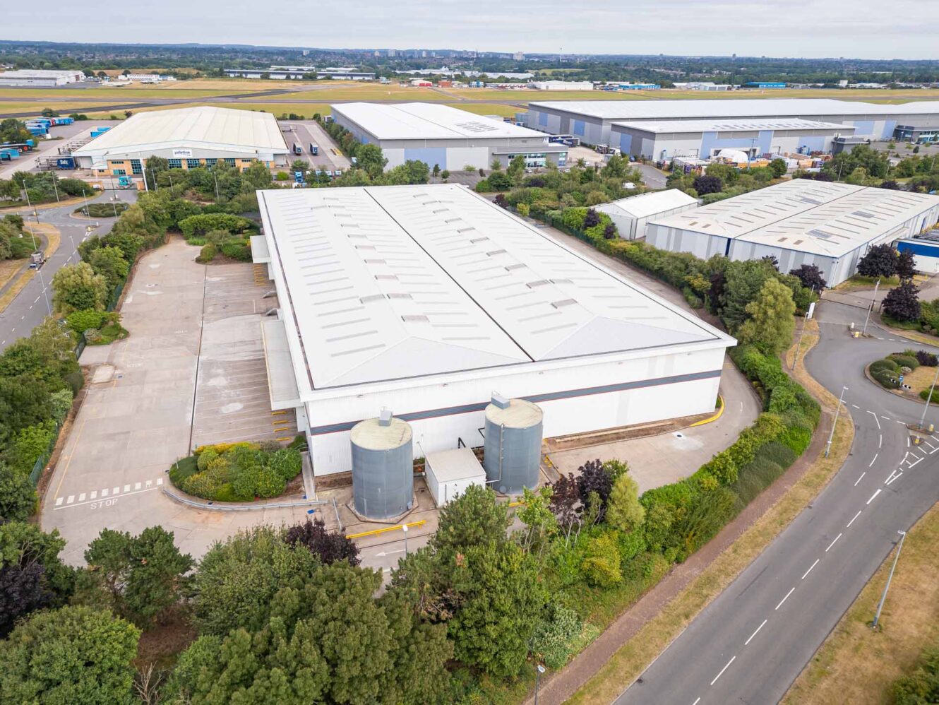 Aerial view of a large industrial warehouse surrounded by trees, empty parking areas, and nearby roads, with other warehouses and industrial buildings in the background.