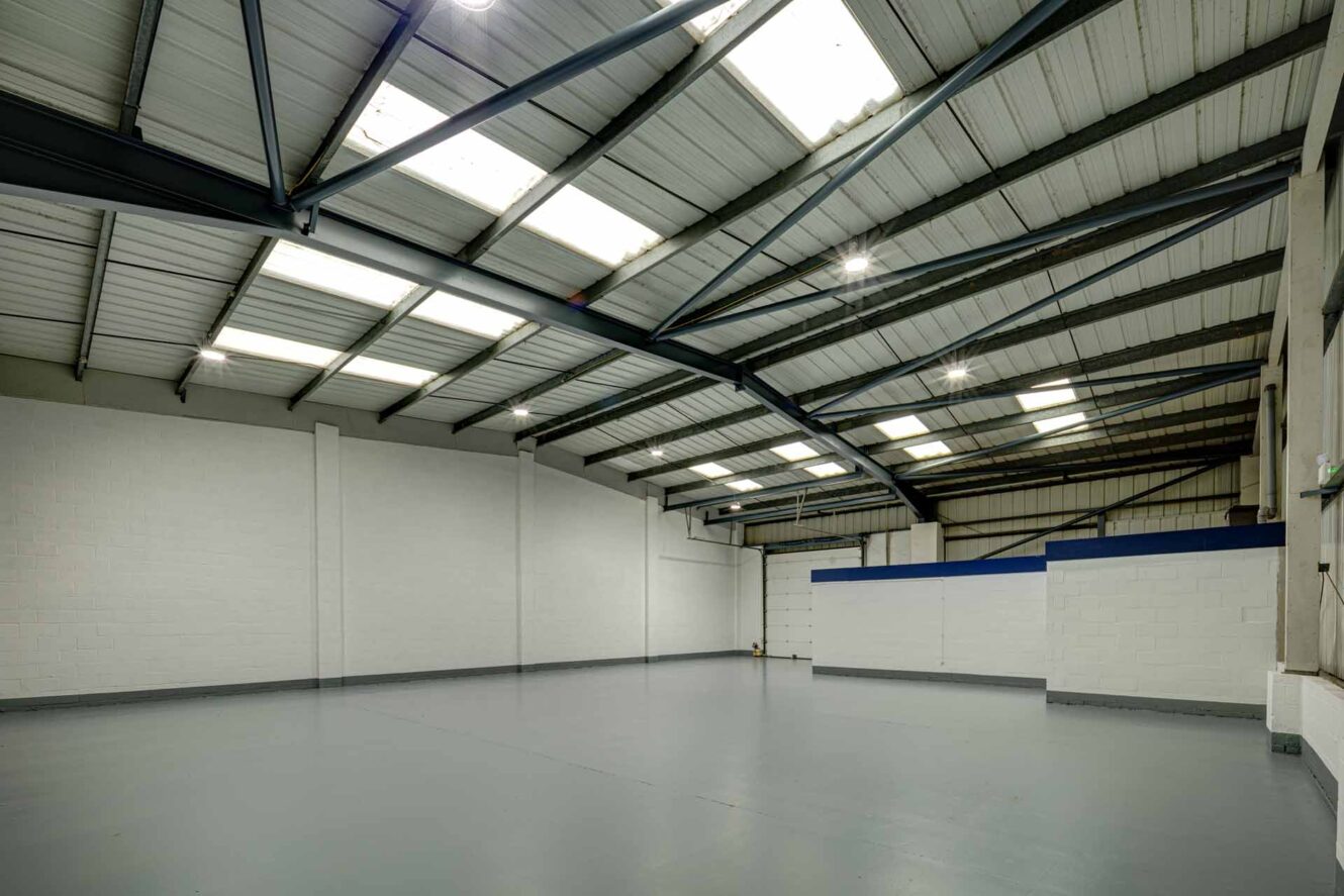 Spacious empty industrial warehouse with grey concrete floor, white painted brick walls, and metal roof with skylights.