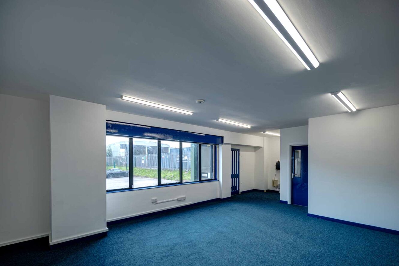 An empty, well-lit office room with blue carpet, white walls, large windows, and blue door frames.