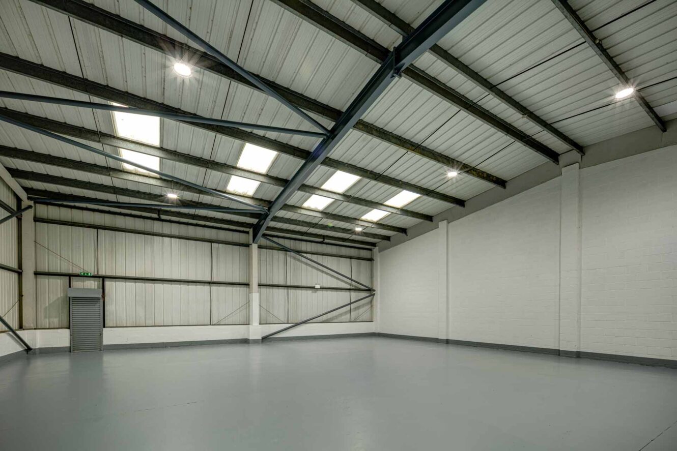 Empty industrial warehouse interior with white walls, grey concrete floor, steel beams, skylights, and fluorescent lighting.