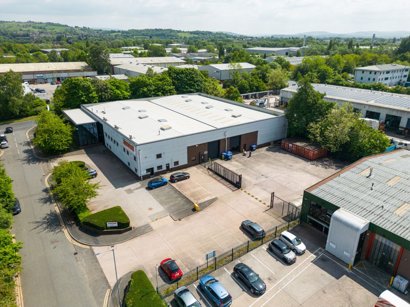 Aerial view of an industrial warehouse with a fenced yard, parked cars, and surrounding trees and buildings on a clear day.