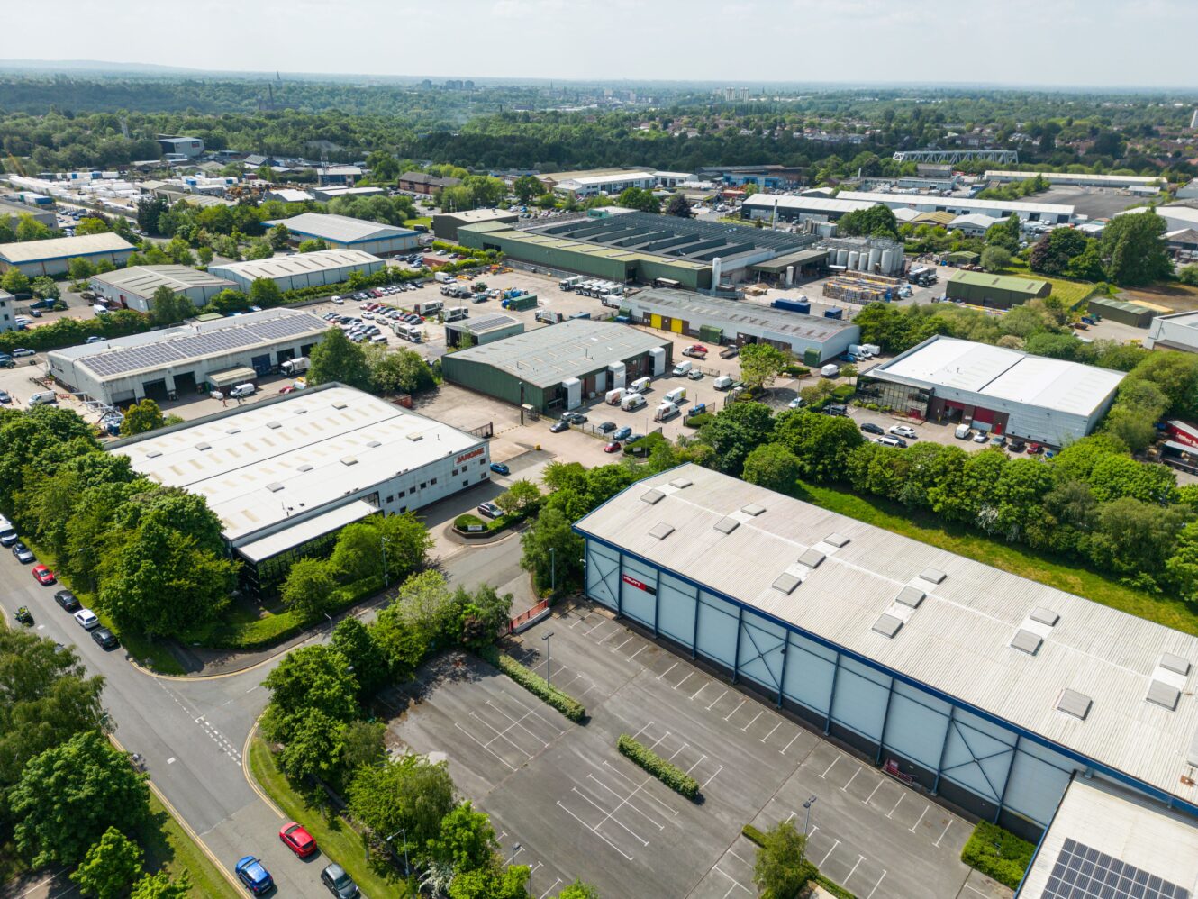Aerial view of an industrial estate with large warehouse buildings, parking lots, several vehicles, and surrounding greenery on a clear day.