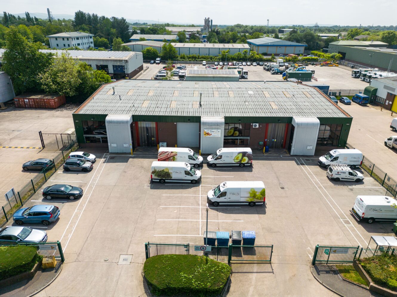 Aerial view of an industrial building with several branded vans and cars parked outside in a gated lot, surrounded by other warehouse structures and greenery.