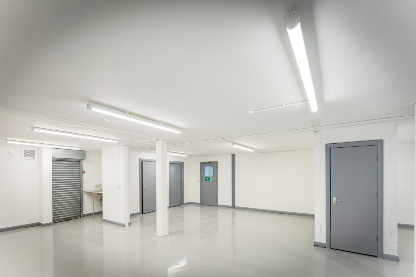 A clean, empty room with gray doors, a metal roller shutter, white walls, overhead fluorescent lights, and a polished gray floor.