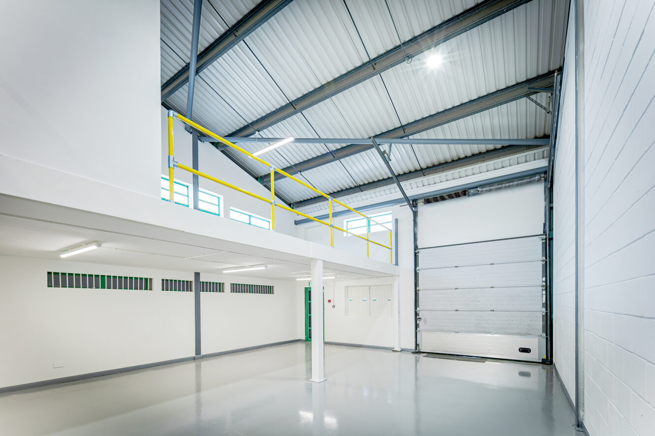 An empty industrial warehouse interior with a mezzanine floor, yellow railings, large overhead door, white walls, and polished concrete floor.