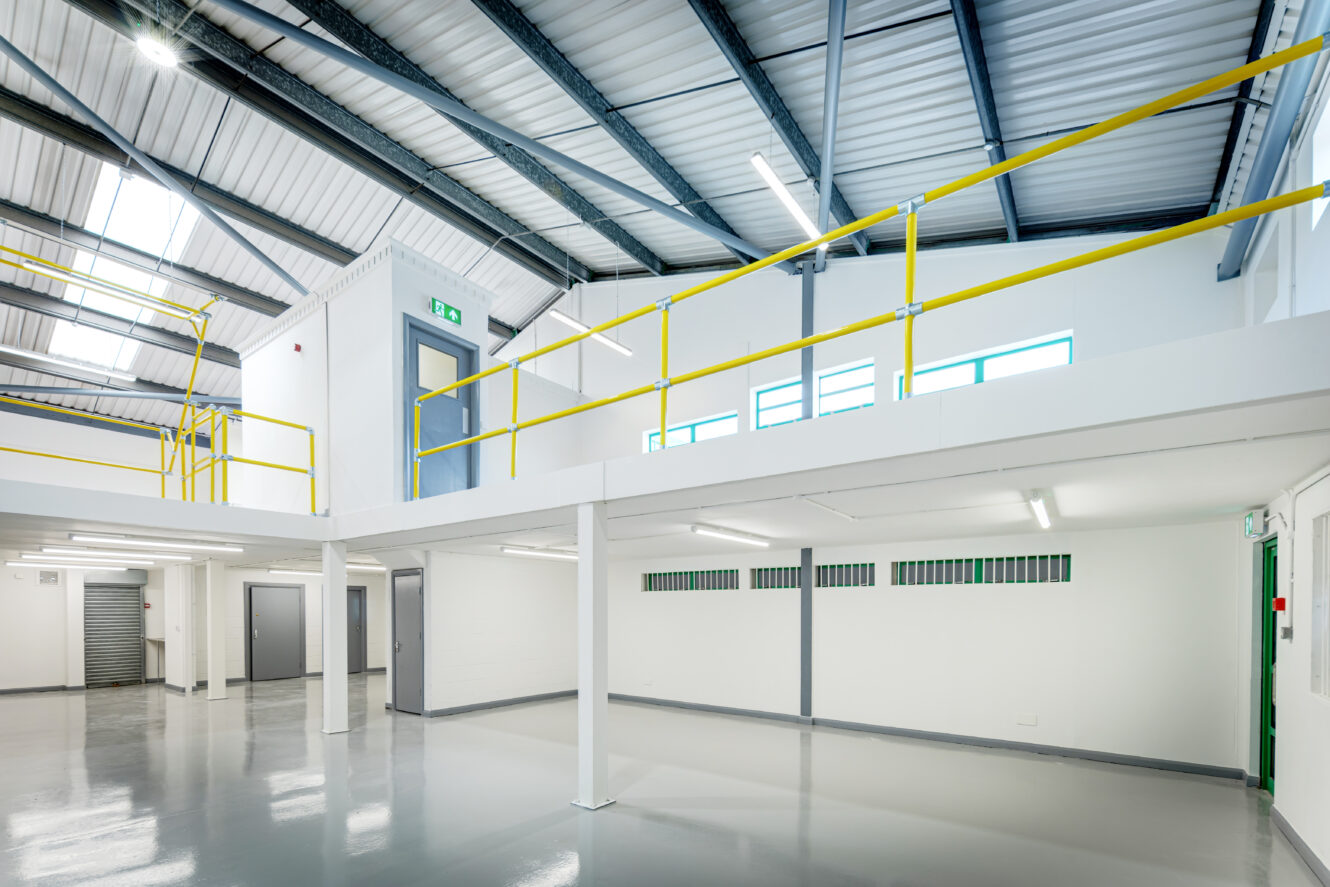 Modern industrial interior with a mezzanine level, white walls, grey floors, yellow railings, and exposed steel beams.