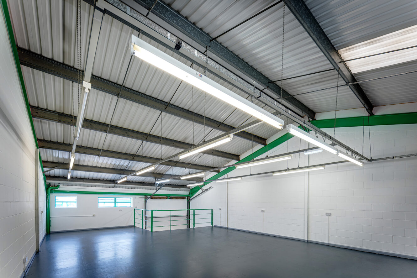 Interior of an empty industrial warehouse with white walls, high ceiling, exposed beams, fluorescent lights, and windows on one side.