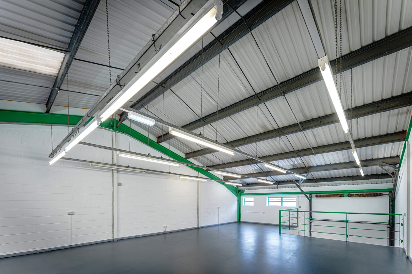 Large, empty industrial warehouse interior with white walls, grey floor, exposed steel beams, fluorescent lighting, and green railings near windows on the right.