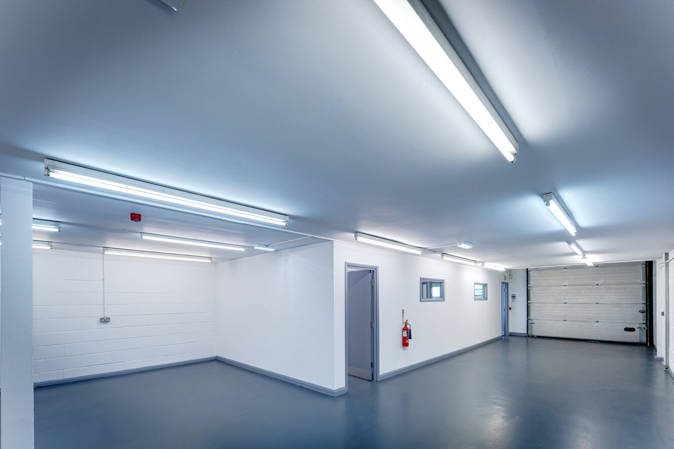 A clean, empty industrial garage or storage space with white walls, grey floors, fluorescent lighting, and a large closed garage door.