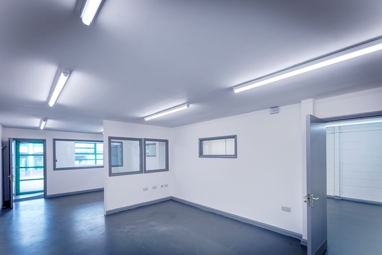 Empty, modern white office space with gray flooring, fluorescent ceiling lights, several windows, and open doors leading to adjacent rooms.
