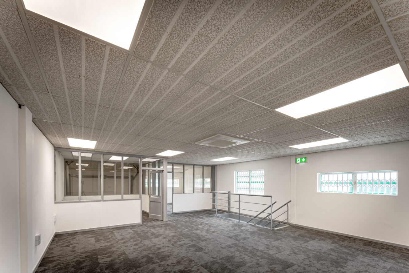 Empty office space with carpeted floor, fluorescent ceiling lights, glass partitioned office, windows with security bars, and a metal railing near a stairwell.