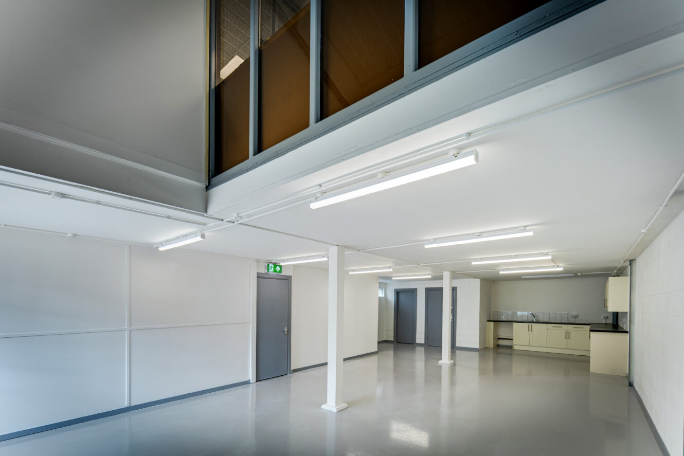 A clean, empty industrial room with white walls, gray doors, support columns, overhead fluorescent lighting, and a kitchenette in the back corner.