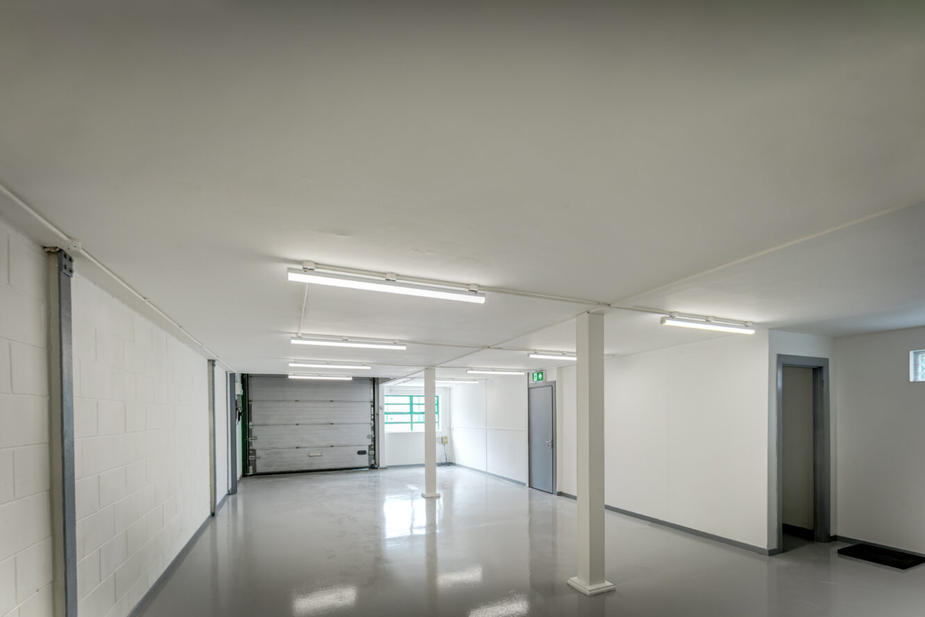 Empty, clean basement with white walls, glossy gray floor, fluorescent ceiling lights, columns, doors, and a small high window.