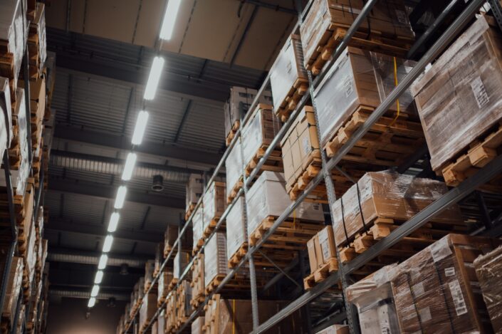 Interior of a warehouse with high shelves holding stacked pallets of boxed goods, seen under bright overhead lighting.