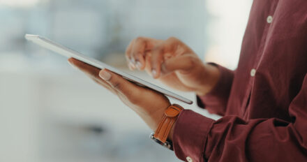 A person wearing a maroon shirt and brown watch uses a tablet, holding it in one hand and touching the screen with the other.