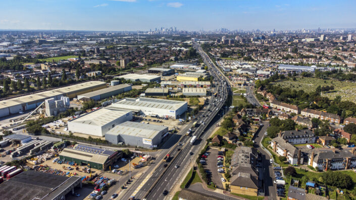 Aerial view of an industrial area with warehouses, houses, and a main road running through, with city skyline visible in the distance under a clear sky.
