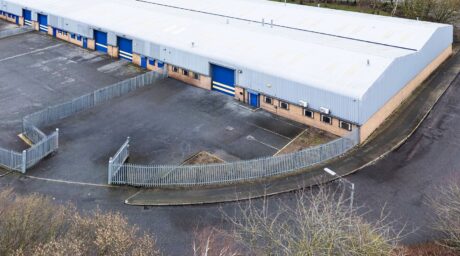 A large industrial warehouse building with blue doors, beige brick lower walls, metal fencing, and an empty paved area in front.