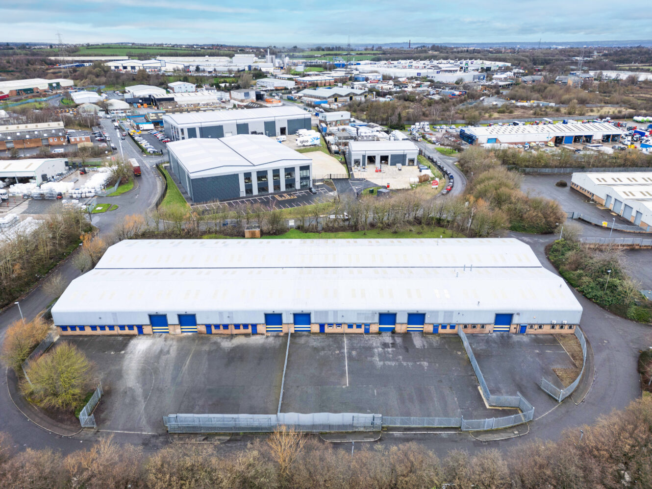 Aerial view of a large industrial warehouse with a white roof and blue doors, surrounded by a fenced lot and other industrial buildings in the background.